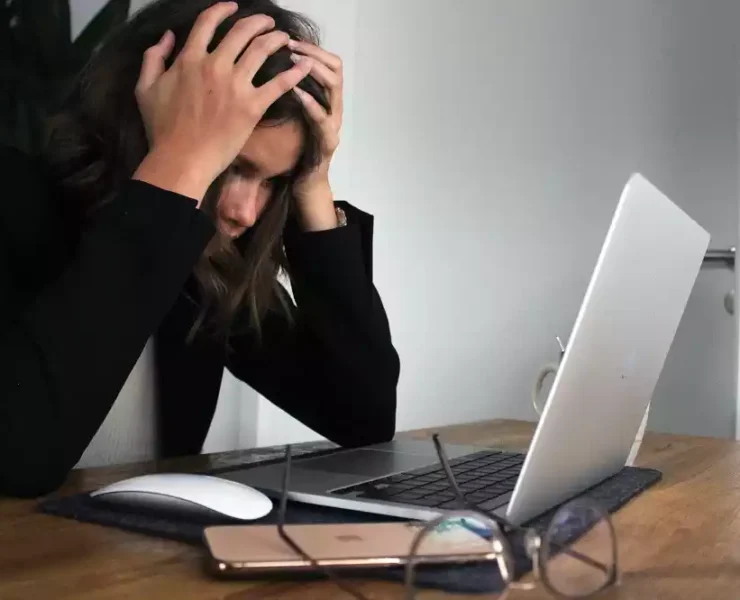 Burned-out woman with hands on head, sitting at table with laptop and wired mouse on it